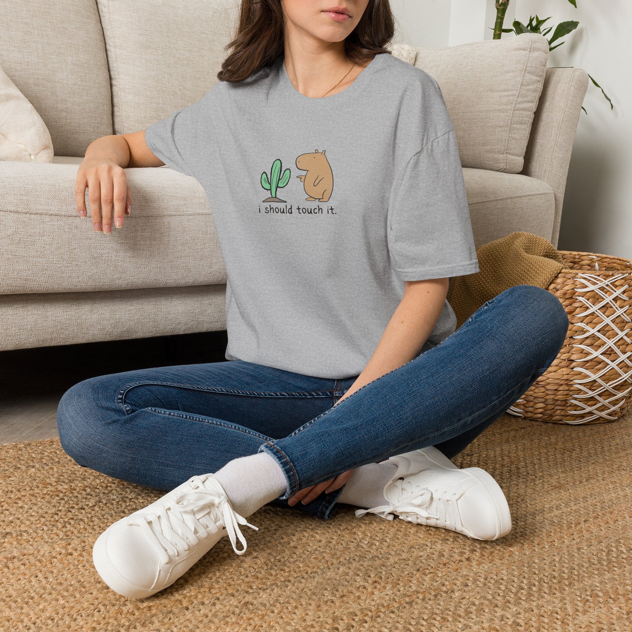 Person wearing a gray short-sleeve crewneck t-shirt featuring a cute graphic of a small green cactus and a brown capybara with the text 'i should touch it.' printed on the chest, displayed against a solid white background sitting on a carpeted floor.