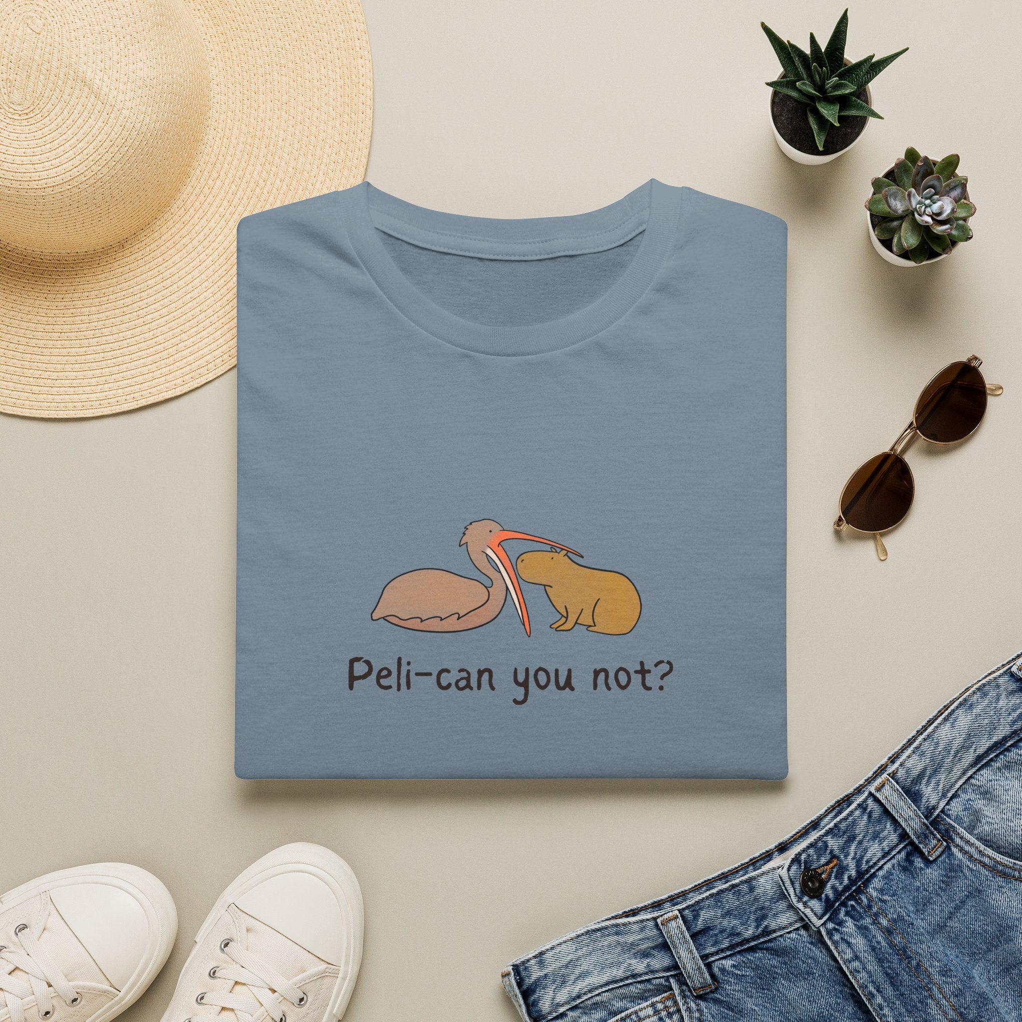 A folded stone blue t-shirt with a pelican attempting to eat a capybara and the humorous text "peli-can you not?" on it ' is shown in a styled flat lay with a straw hat, white sneakers, blue jeans, sunglasses, and small potted succulents on a light beige background.