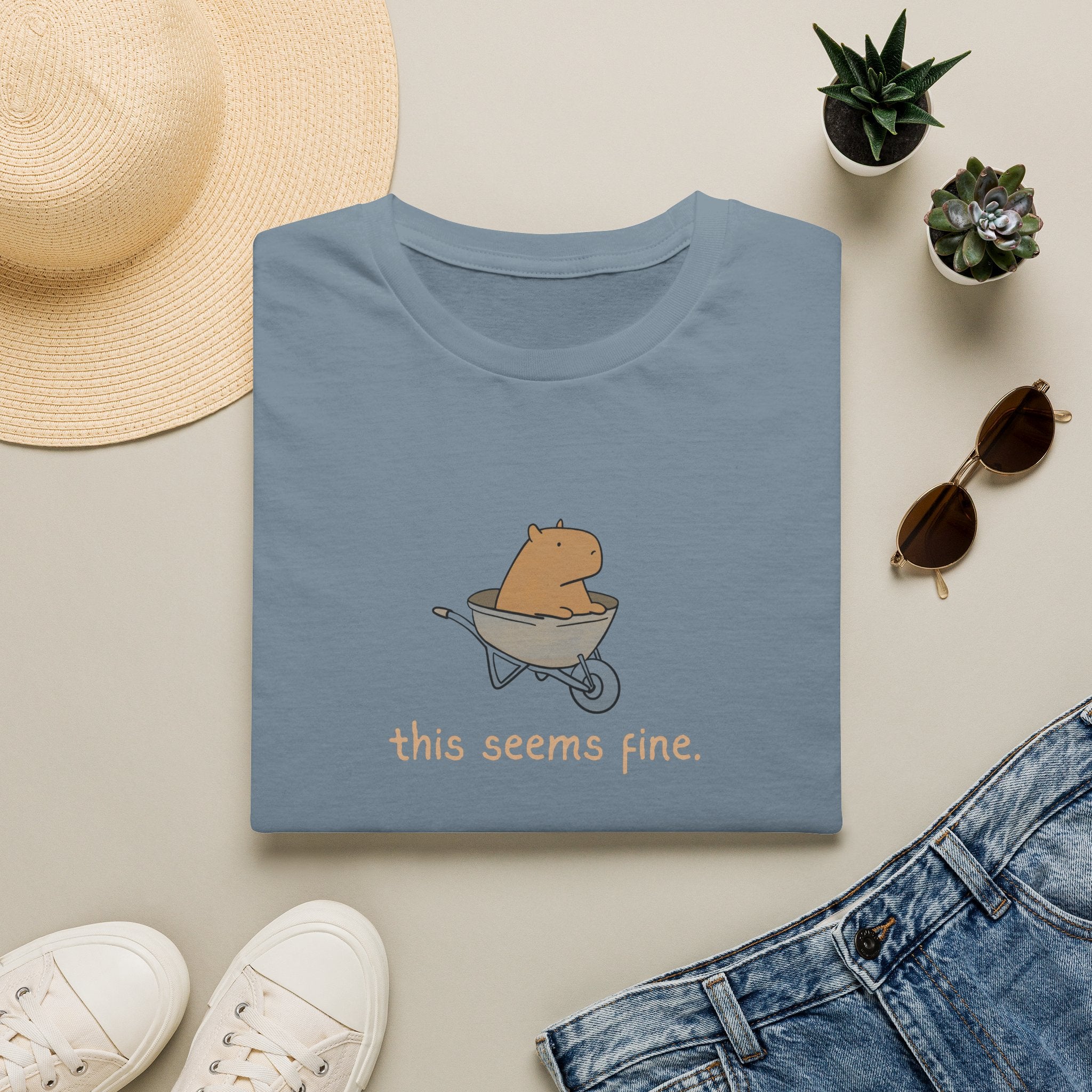 Folded blue tshirt with a capybara in a wheelbarrow and the humorous text "this seems fine." on a light surface with a hat, sunglasses, and jeans.