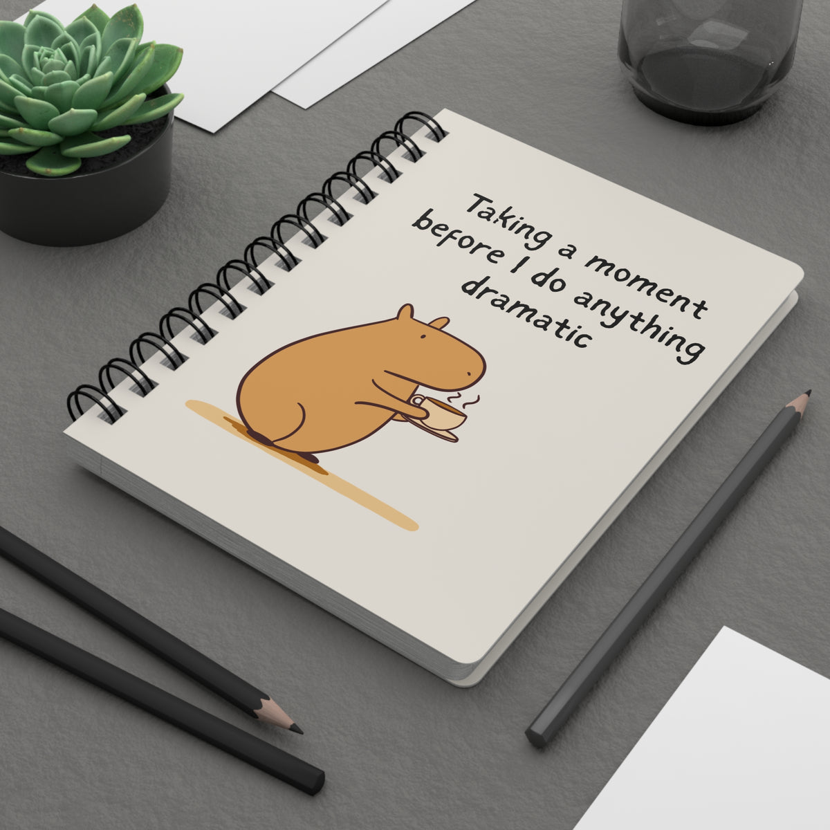 A spiral-bound notebook lying on a desk with a cream cover featuring a capybara holding a steaming coffee cup, with the words 'Taking a moment before I do anything dramatic' above.