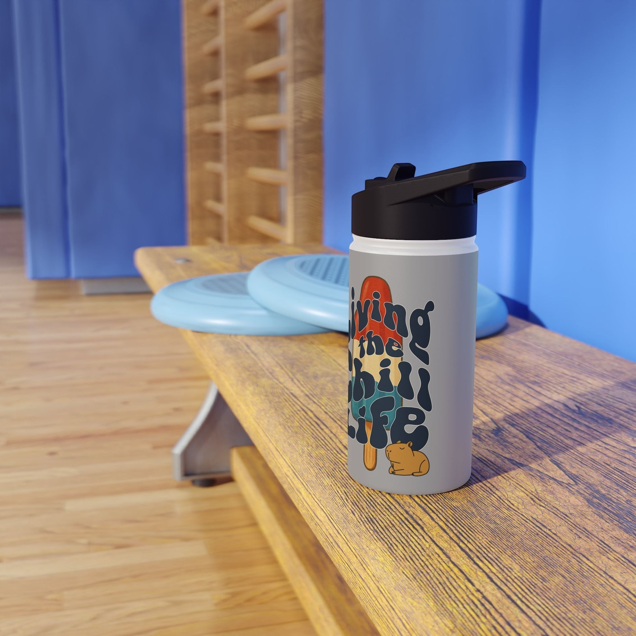 Capy Life insulated kids water bottle with retro popsicle design and capybara illustration on a wooden gym bench.