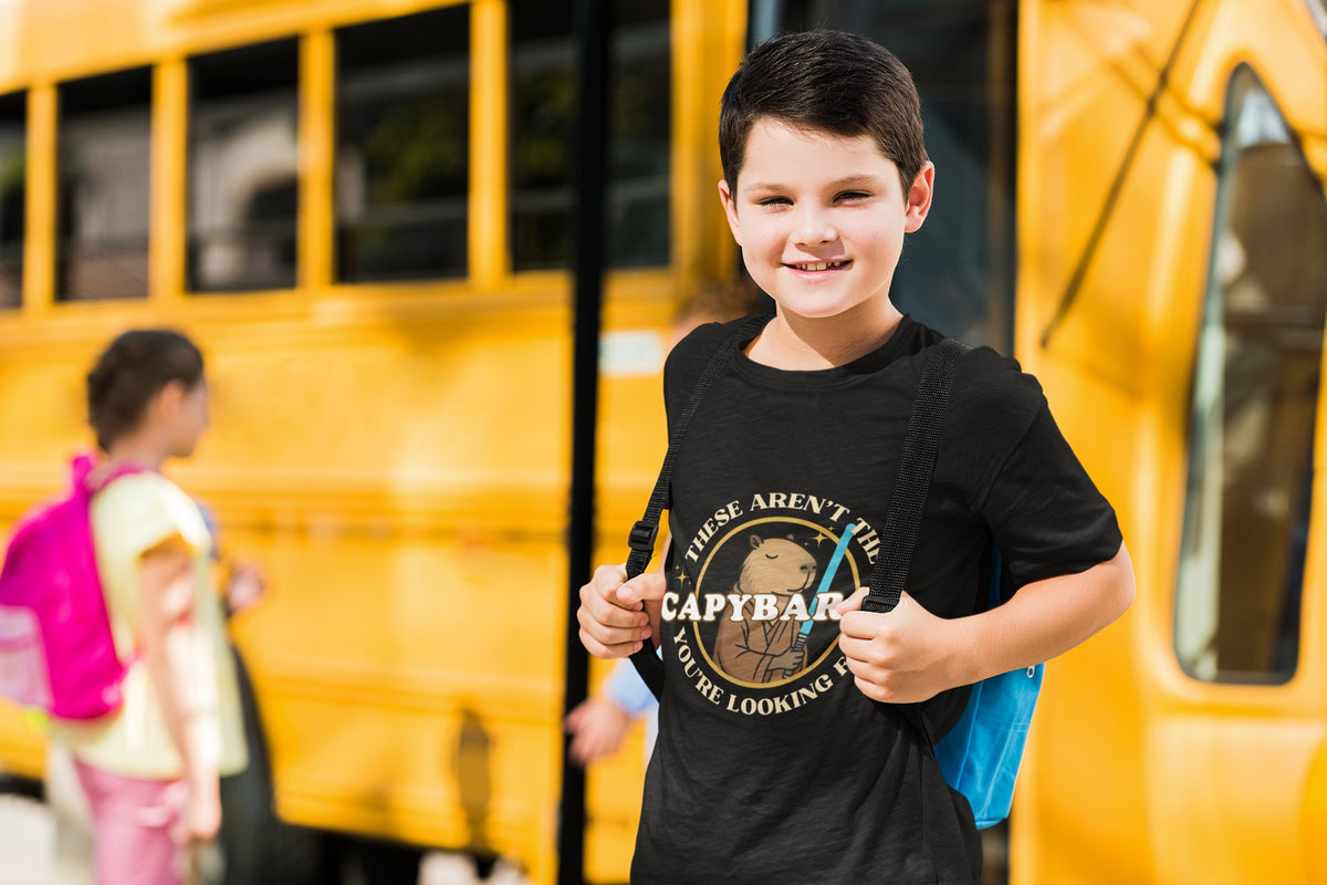 Capy Life youth wearing a black cotton t-shirt with a Jedi capybara graphic standing in front of a yellow school bus