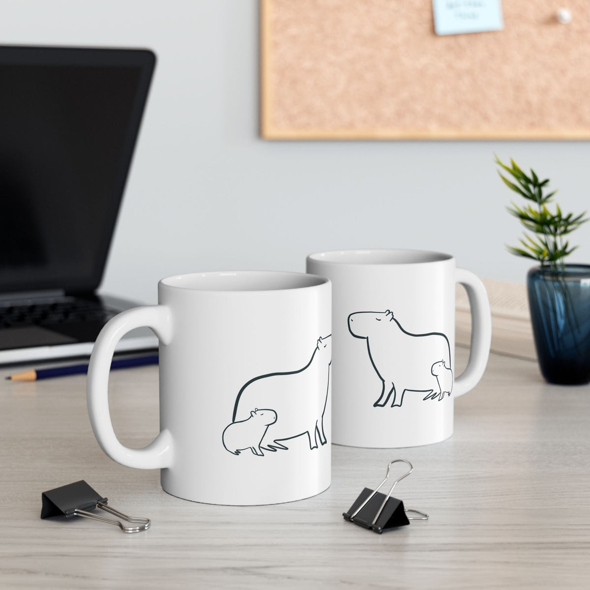 Capy Life white ceramic mug featuring a minimalist black line drawing of a mother capybara and her baby on a desk.