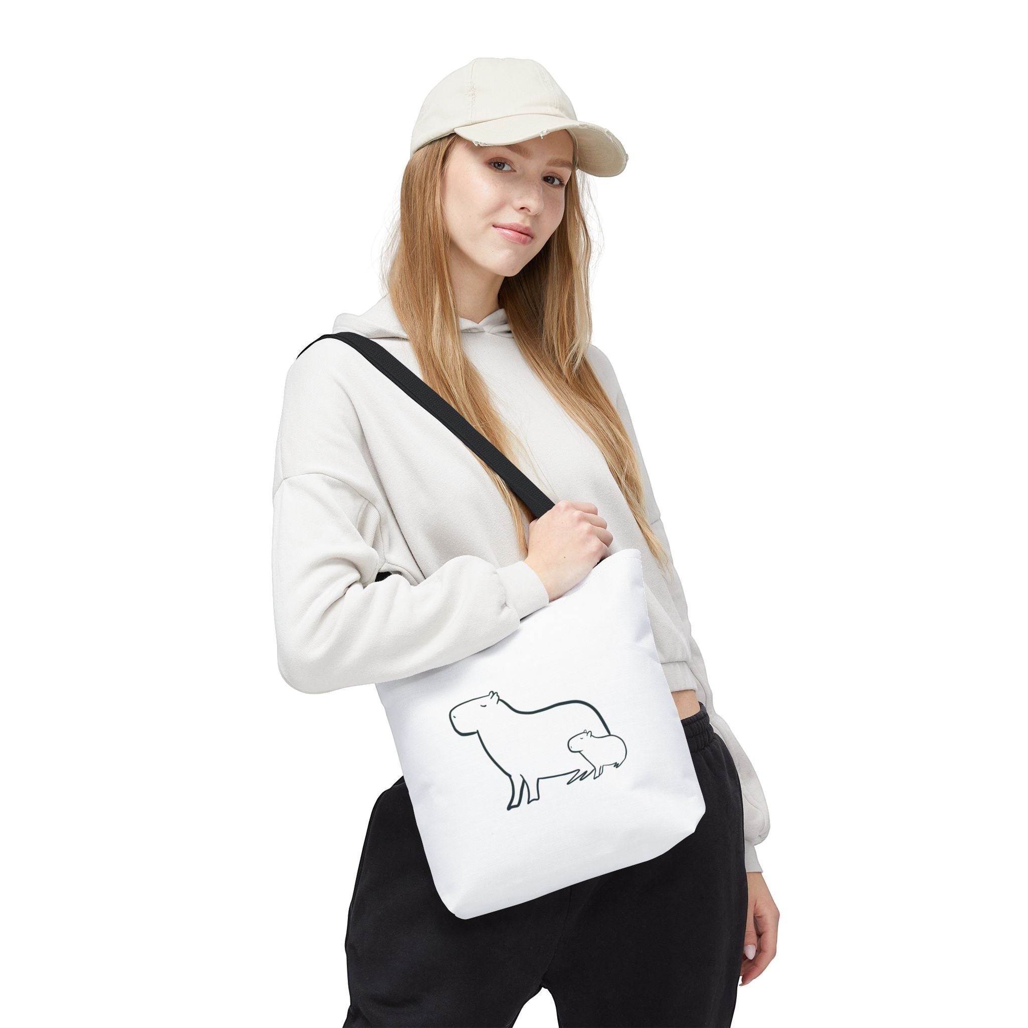 A woman wearing a beige hoodie and cap carries a white Capy Life minimalist tote bag with a capybara line art print.