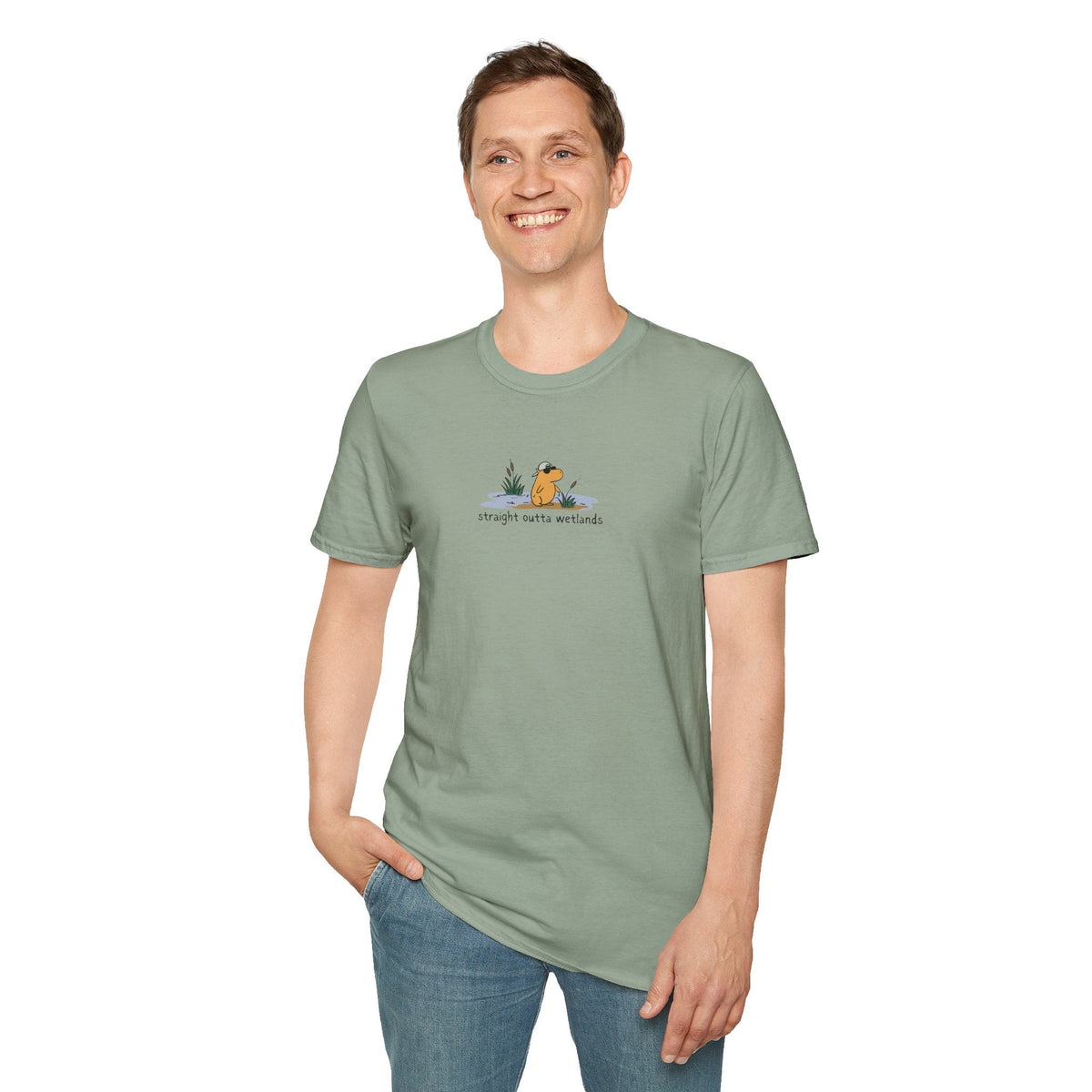 Man wearing a sage green Capy Life softstyle t-shirt featuring a small capybara in a backwards hat graphic.