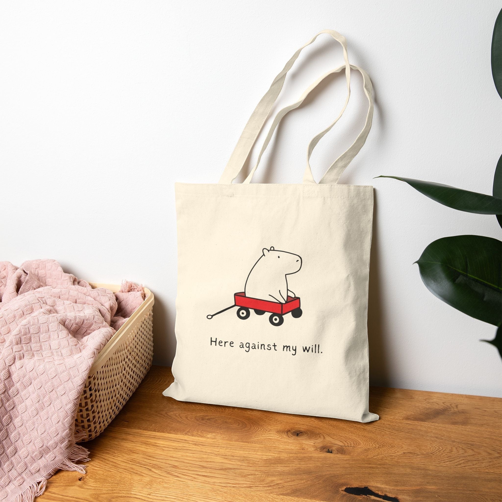 Capy Life natural canvas tote bag featuring a cute capybara in a red wagon with the text Here against my will.