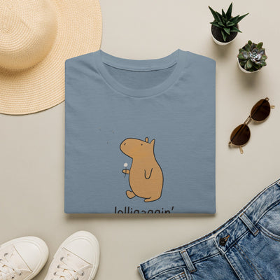 A folded dusty blue t-shirt with a cartoon capybara holding a lollipop and the text 'lolligaggin' underneath, styled in a flat lay with a straw hat, white sneakers, sunglasses, a pair of blue jeans, and small potted succulents on a light beige background.