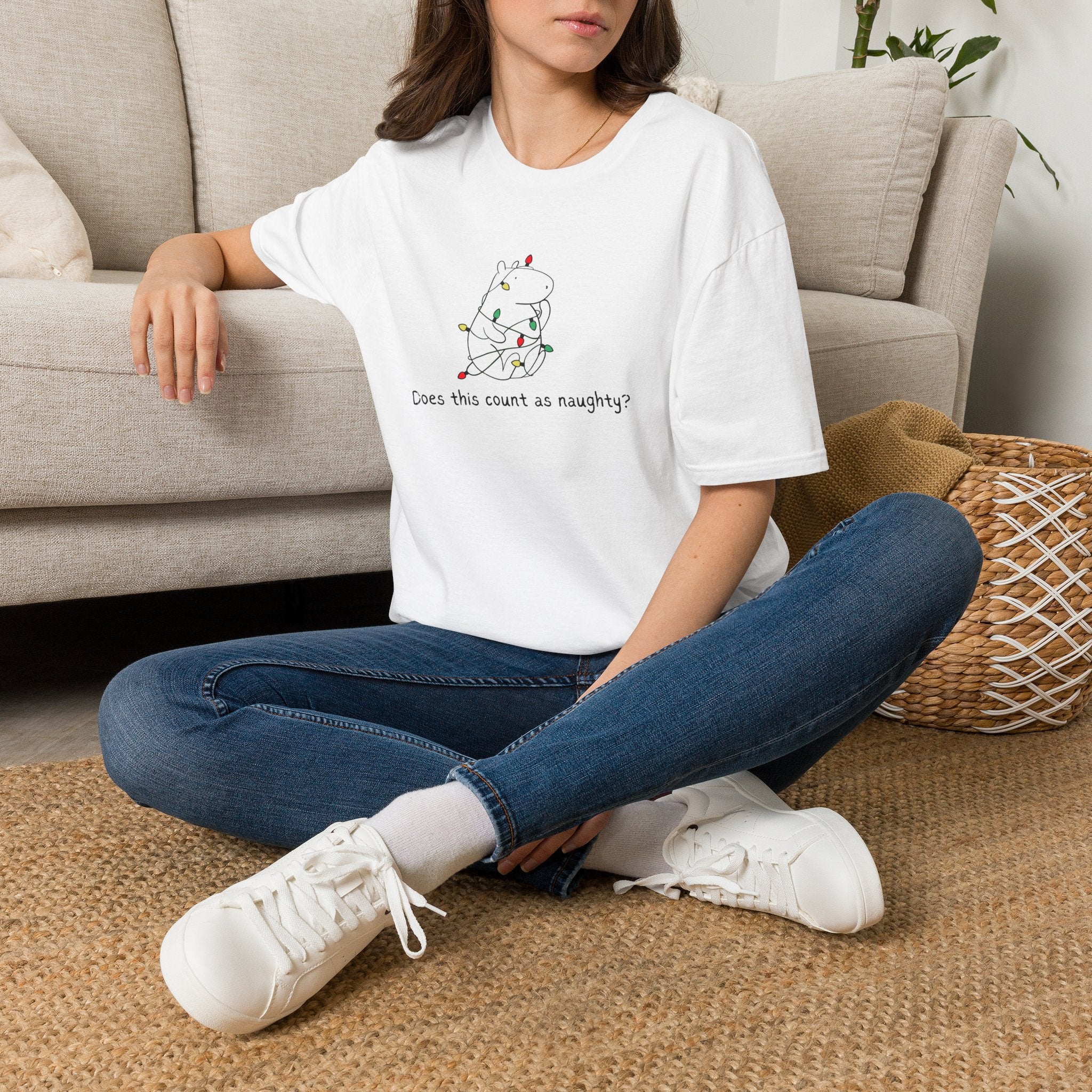 Person sitting on the floor wearing a white  t-shirt with a capybara tangled in christmas lights and the humorous phrase "Does this count as naughty?" blue jeans, and white sneakers.