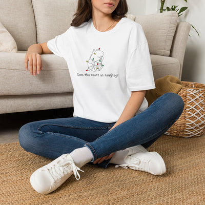 Person sitting on the floor wearing a white  t-shirt with a capybara tangled in christmas lights and the humorous phrase "Does this count as naughty?" blue jeans, and white sneakers.