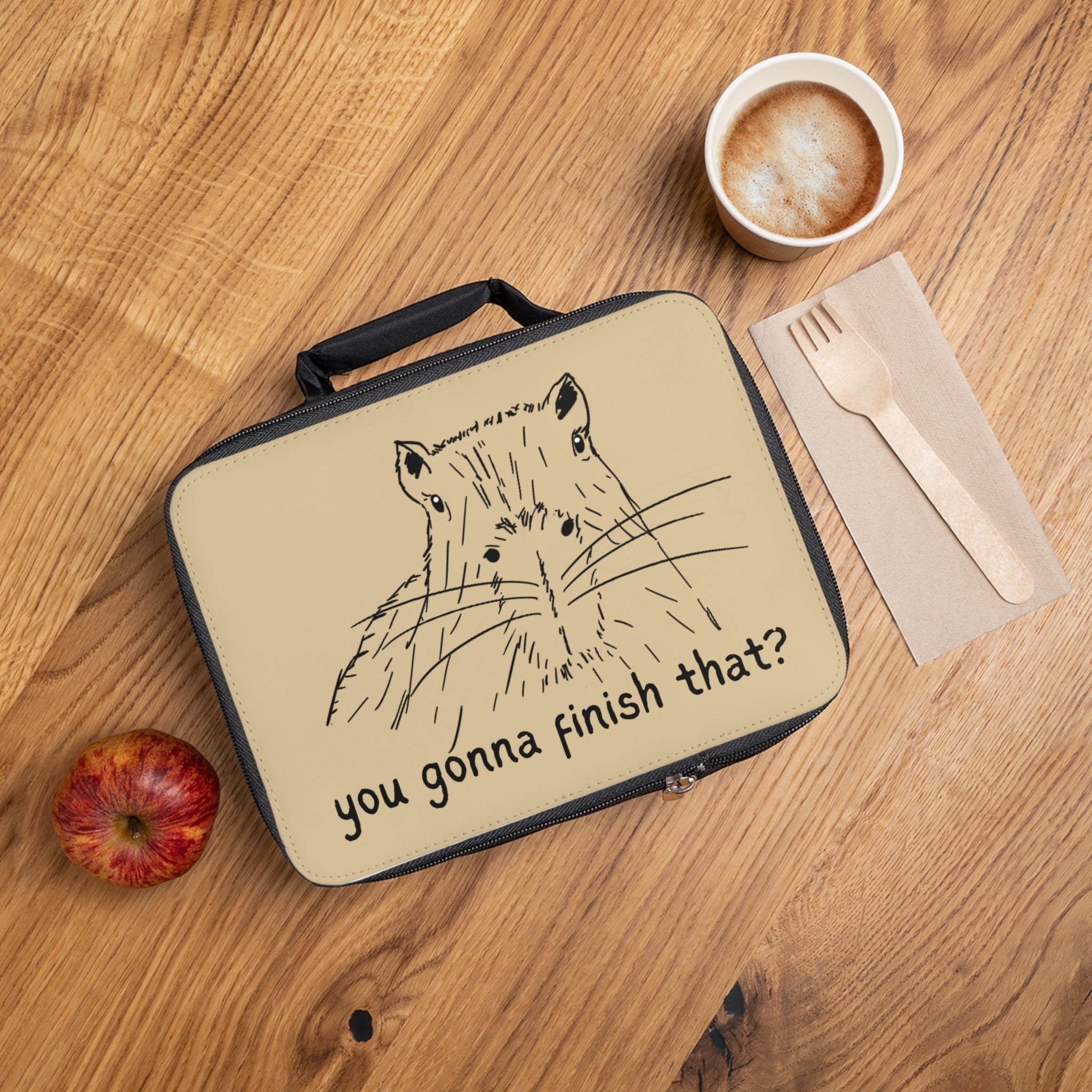 A beige lunch bag with a black handle and zipper, featuring a black line art illustration of a capybara and the text 'you gonna finish that?' on the front. The bag is shown in a flat-lay arrangement on a wooden table next to a cup of coffee, a wooden fork on a napkin, and an apple.