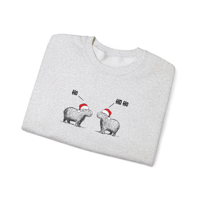 A light heather gray crewneck sweatshirt folded flat, featuring a graphic of two capybaras wearing red Santa hats with 'Ho' and 'Ho Ho' text above them, set against a plain white background.