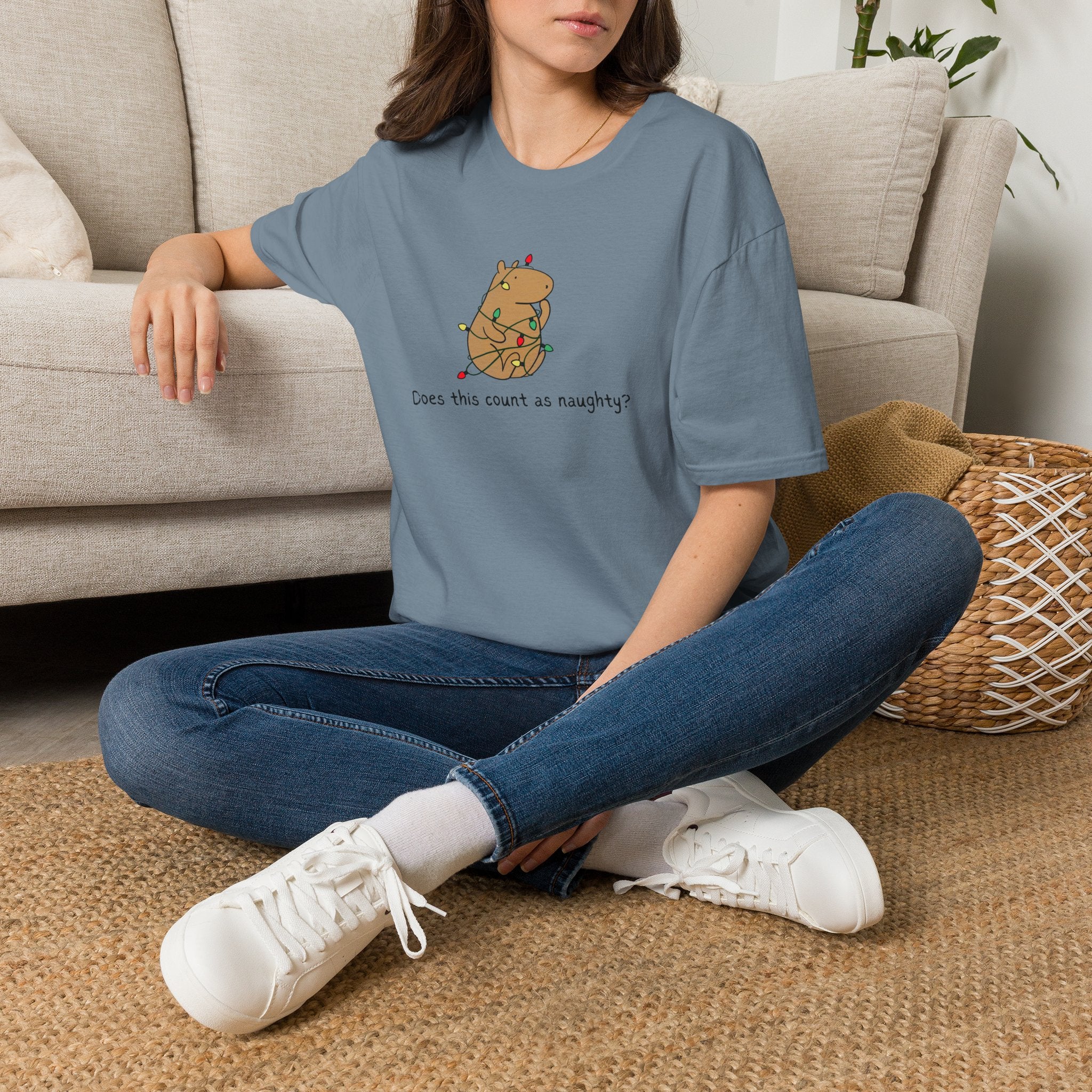 A woman sitting cross-legged on a woven rug in front of a beige sofa, wearing a slate blue  t-shirt with a capybara tangled in christmas lights and the humorous phrase "Does this count as naughty?"