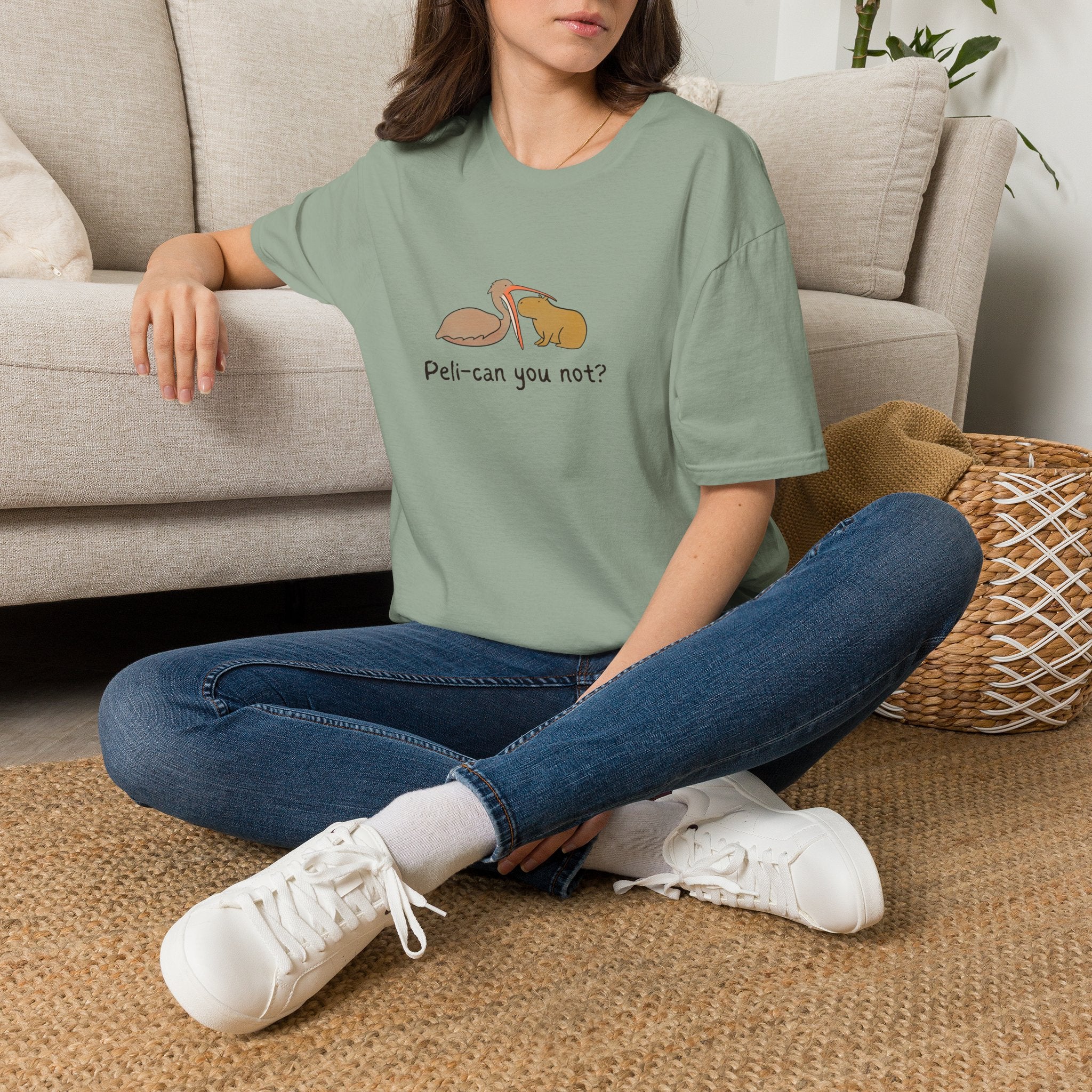 Person sitting on a carpeted floor wearing a green t-shirt with a hand drawn pelican and capybara with humorous text and jeans.