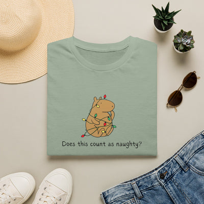 Folded green  t-shirt with a capybara tangled in christmas lights and the humorous phrase "Does this count as naughty?" surrounded by a hat, sunglasses, shoes, and plants on a light surface.