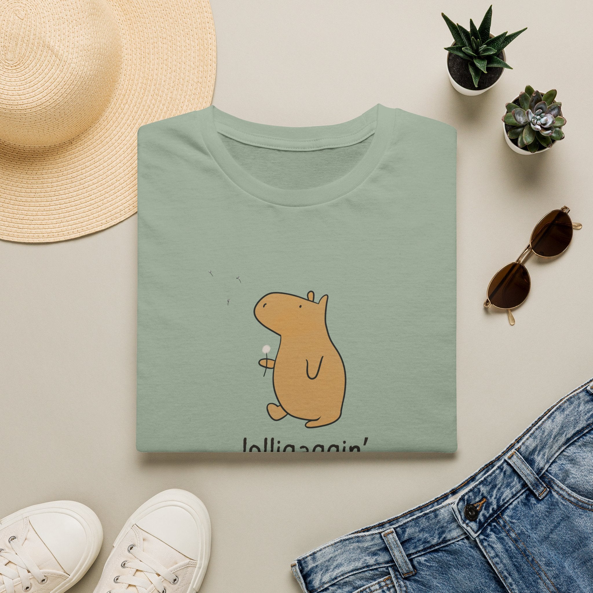 A folded sage green t-shirt with a cute cartoon capybara holding a flower, styled in a flat lay with a straw hat, white sneakers, sunglasses, a pair of blue jeans, and small potted succulents on a light beige background.