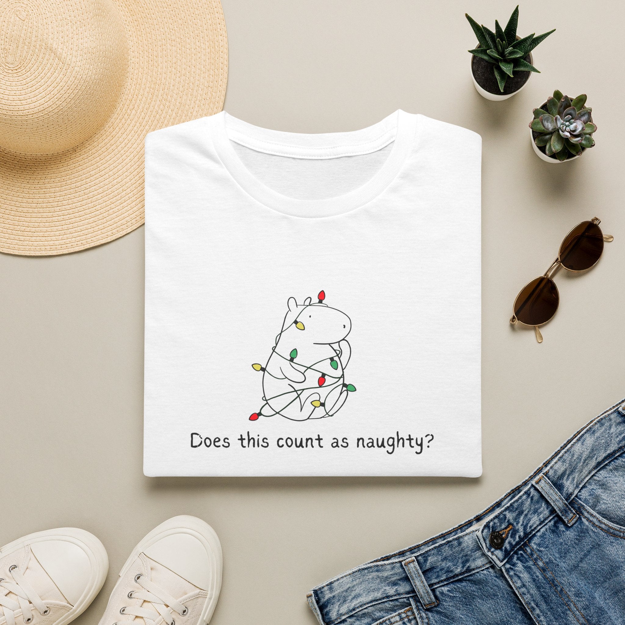 white  t-shirt with a capybara tangled in christmas lights and the humorous phrase "Does this count as naughty?" on a beige surface with casual items like a hat, sunglasses, and shoes.