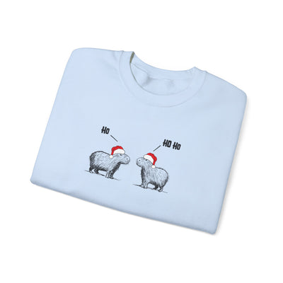 A light blue crewneck sweatshirt folded flat, featuring a graphic of two capywars wearing red Santa hats with 'Ho' and 'Ho Ho' text above them, set against a plain white background.