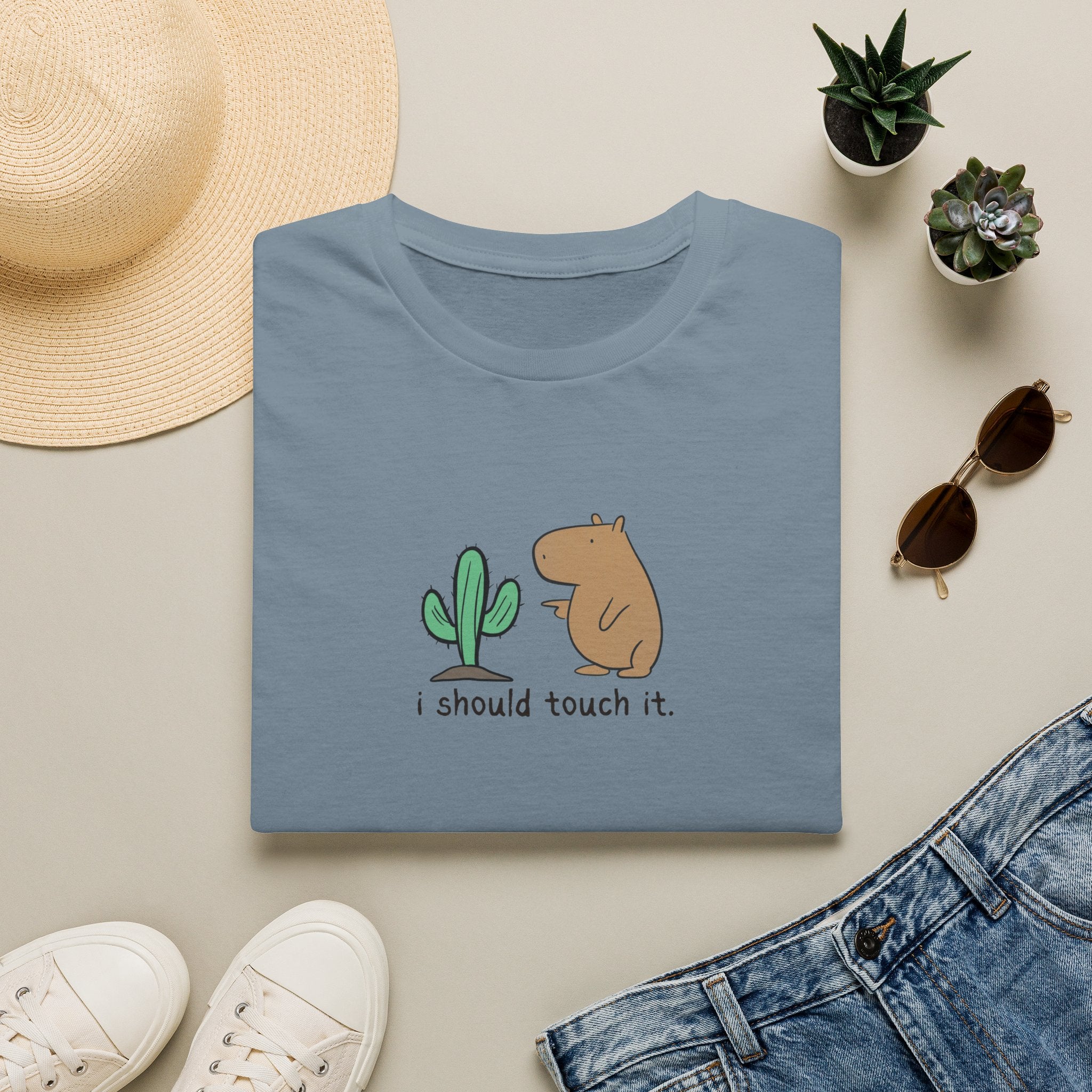 A folded dusty blue t-shirt with a cute cartoon graphic of a capybara and a cactus and the text 'i should touch it.' displayed in a styled flat-lay with a straw hat, white sneakers, sunglasses, and denim jeans on a light beige background.