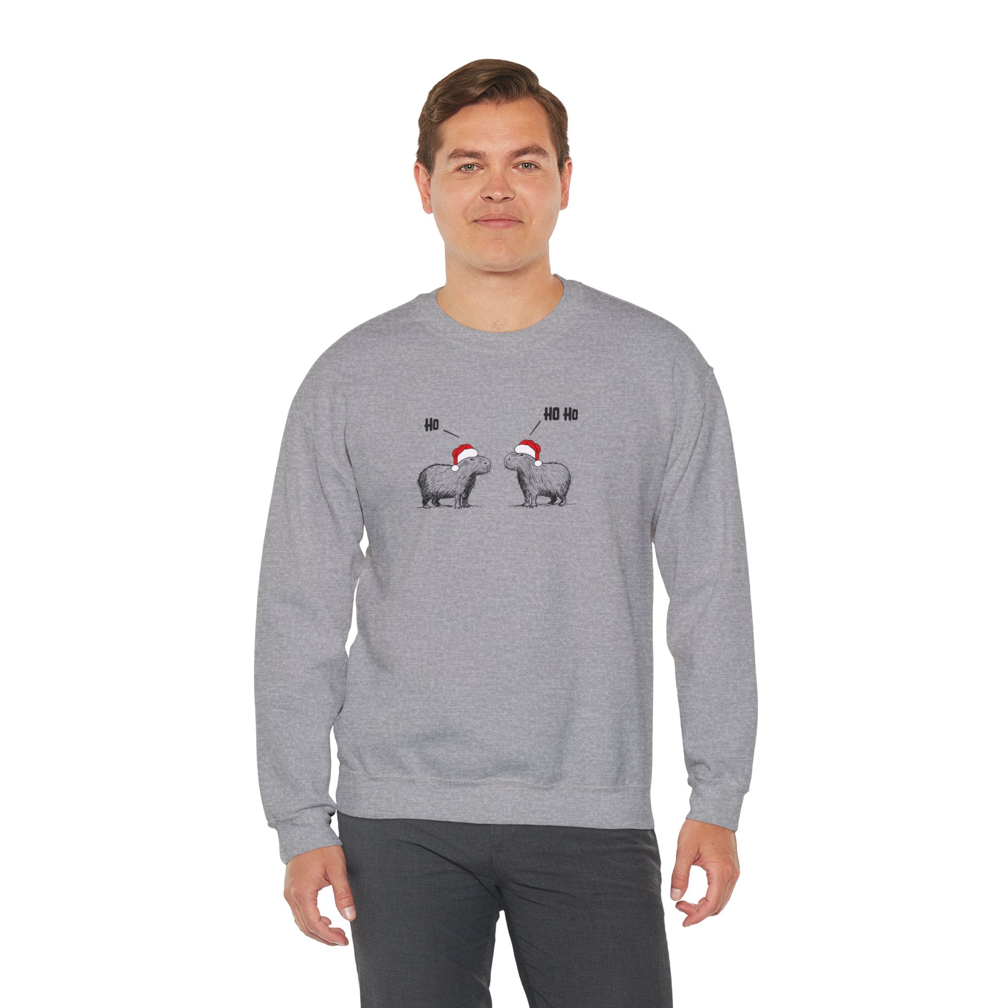 A man wearing a heather gray crewneck sweatshirt featuring a graphic of two capybaras wearing red Santa hats facing each other with 'HO HO HO' text above them