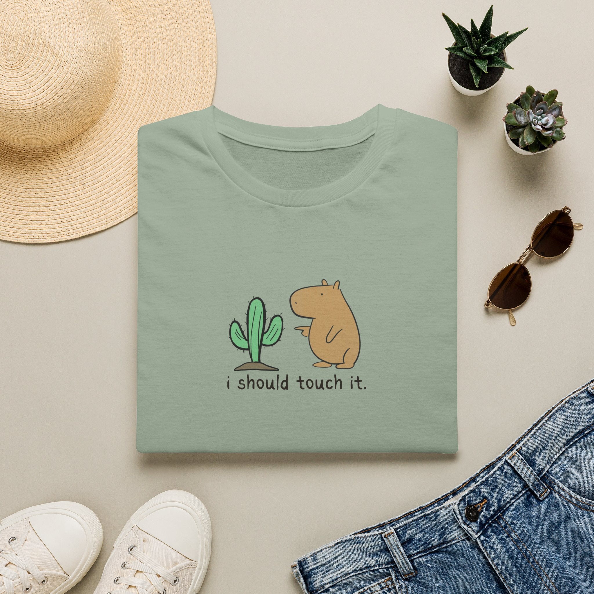 A folded sage green t-shirt with a cute cartoon graphic of a cactus and a capybara and the text 'i should touch it.' displayed in a styled flat-lay with a straw hat, white sneakers, blue jeans, and small potted succulents on a light beige background.