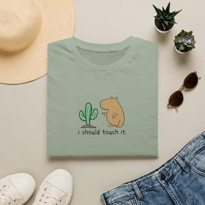 A folded sage green t-shirt with a cute cartoon graphic of a cactus and a capybara and the text 'i should touch it.' displayed in a styled flat-lay with a straw hat, white sneakers, blue jeans, and small potted succulents on a light beige background.