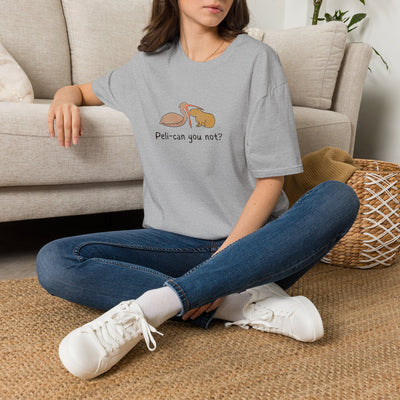 Person sitting on a carpeted floor wearing a gray pelican and capybara punny t-shirt with a graphic design and text, blue jeans, and white sneakers.