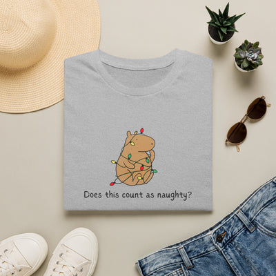A folded light gray T-shirt with a cute cartoon capybara wrapped in Christmas lights and the text 'Does this count as naughty?' is shown in a styled flat-lay with a straw hat, white sneakers, blue jeans, sunglasses, and small potted succulents on a beige background.
