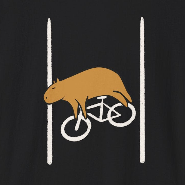 A black short-sleeve crewneck t-shirt featuring a graphic of a brown capybara riding a white bicycle between two vertical white lines on the chest, displayed against a solid white background.