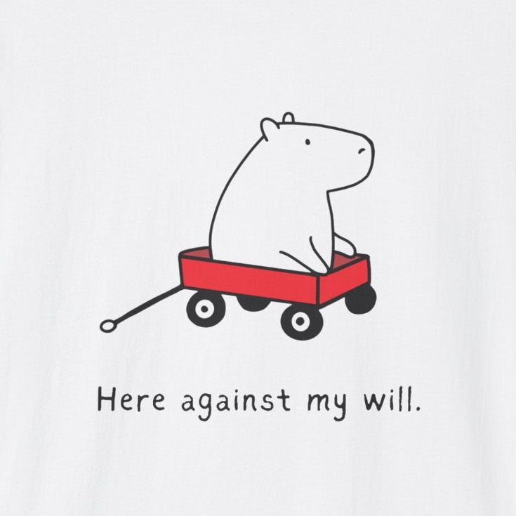 A white short-sleeve crewneck t-shirt featuring a cartoon graphic of a white capybara pulling a red wagon with the text 'Here against my will.' printed below it.