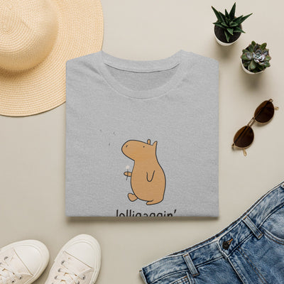 A folded light gray T-shirt with a cartoon capybara graphic in tan and brown, styled in a flat lay with a straw hat, white sneakers, sunglasses, small potted plants, and blue jeans on a light beige background.