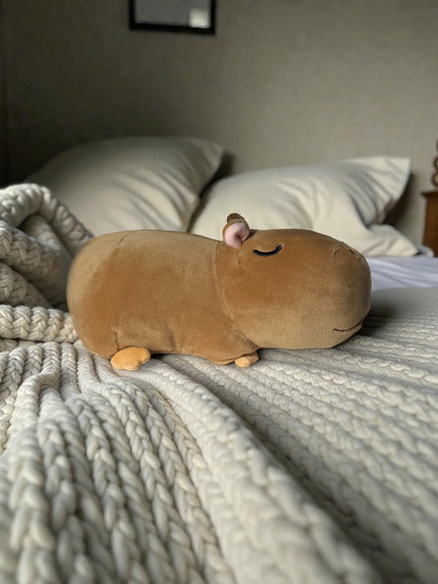 Gene capybara stuffed animal resting on cozy bed with knit blanket and pillows
