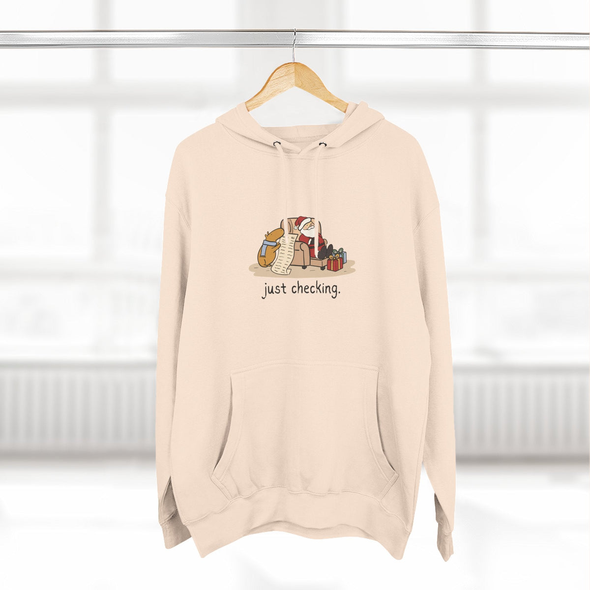 Capy Life beige fleece hoodie featuring a cute capybara checking Santa's naughty-or-nice list, with a kangaroo pocket.