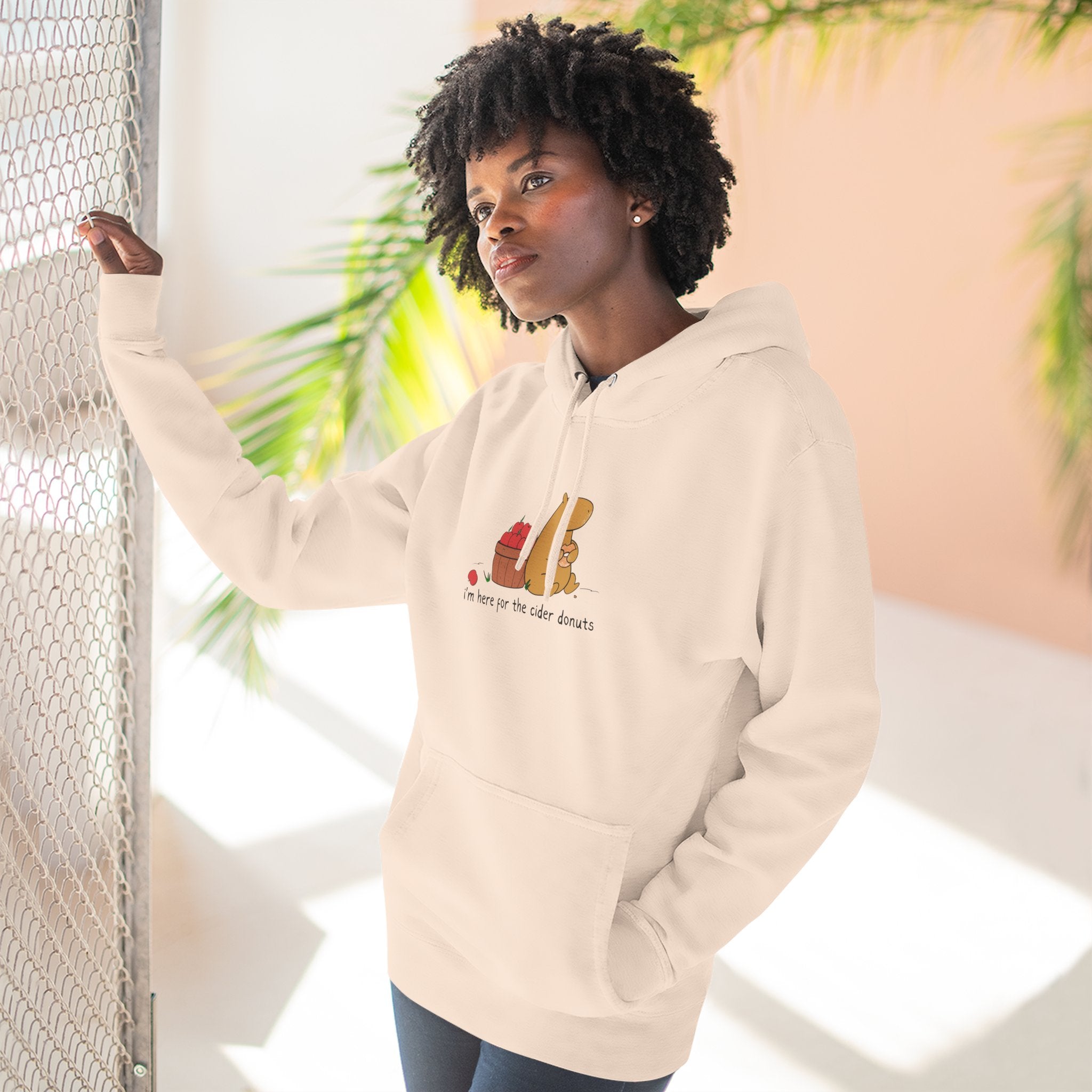 A woman wearing a Capy Life beige fleece hoodie with a capybara eating a cider donut illustration on the chest.
