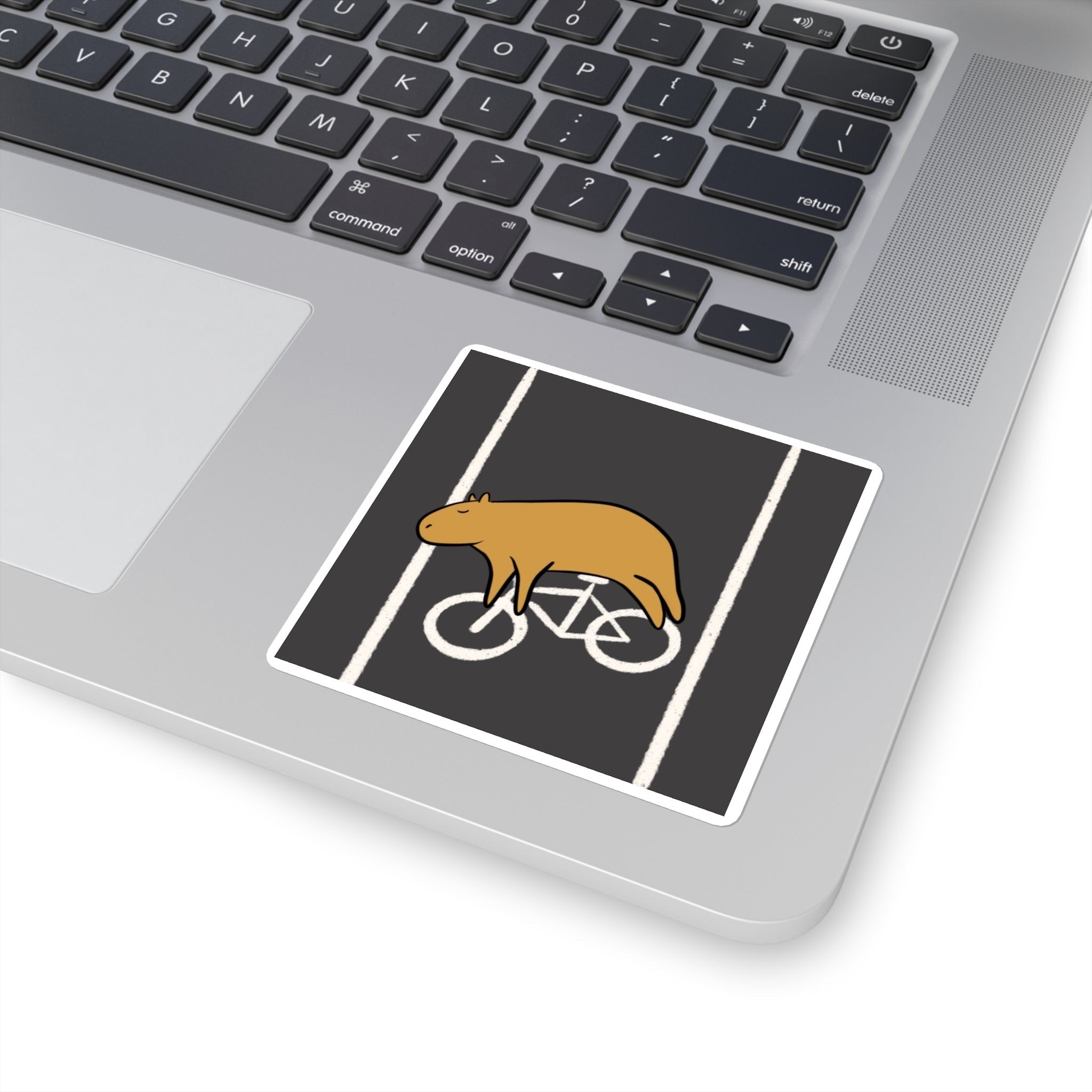 Capy Life vinyl sticker of a relaxed capybara lying in a bike lane, applied to a silver laptop surface.
