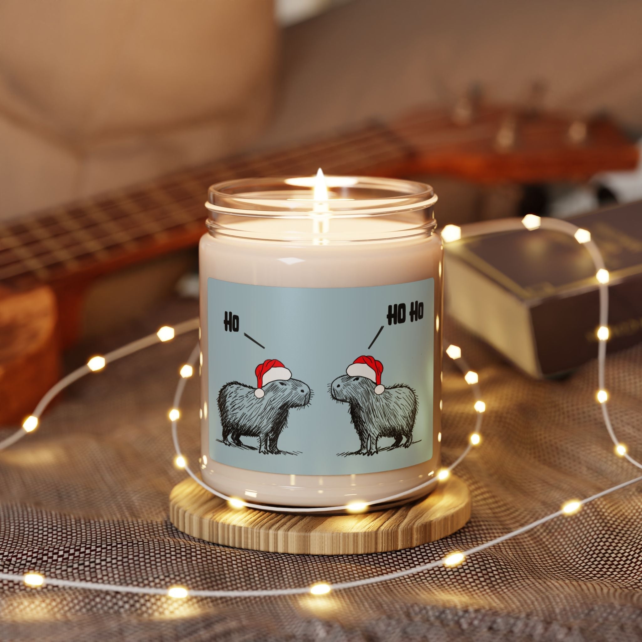 Capy Life Christmas candle featuring two capybaras in Santa hats on a blue label with Ho Ho text, lit on a coaster.