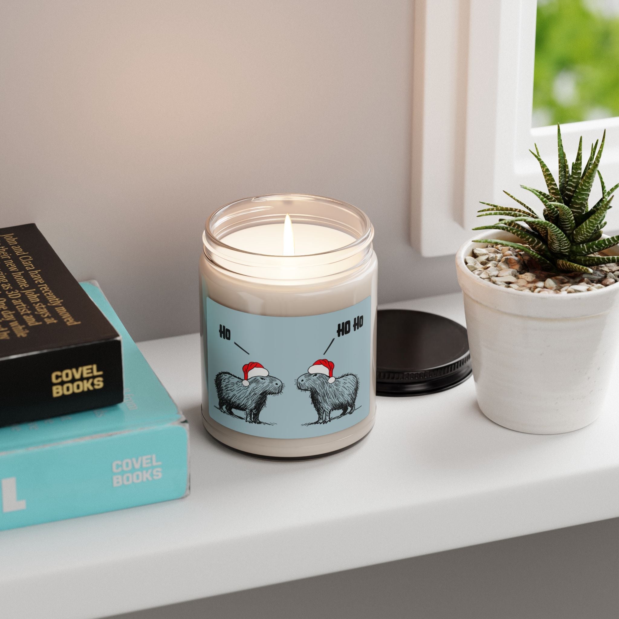 Capy Life Christmas candle featuring two capybaras in Santa hats on a blue label, sitting on a windowsill.