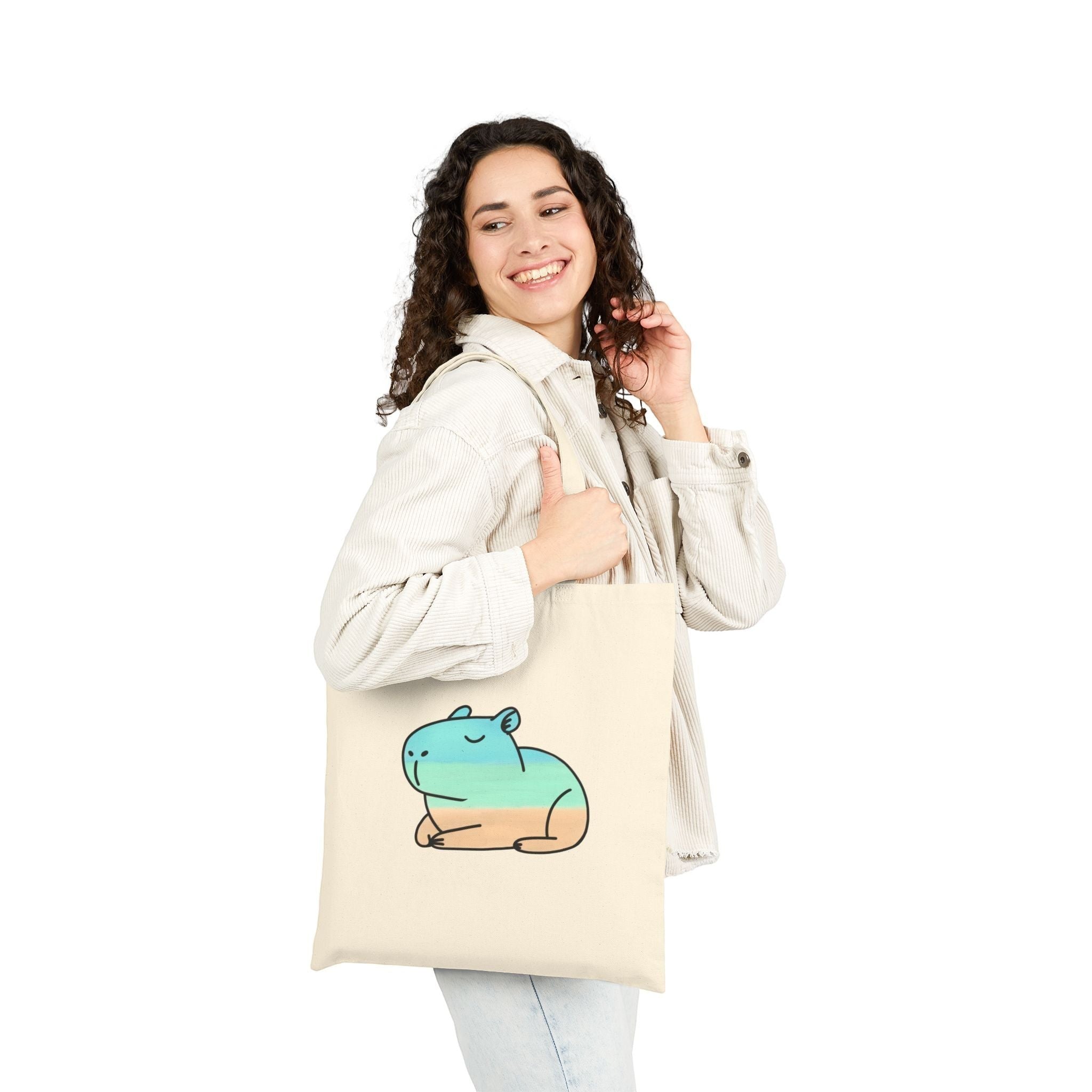 A woman smiling while carrying a Capy Life natural cotton canvas tote bag featuring a coastal capybara illustration.