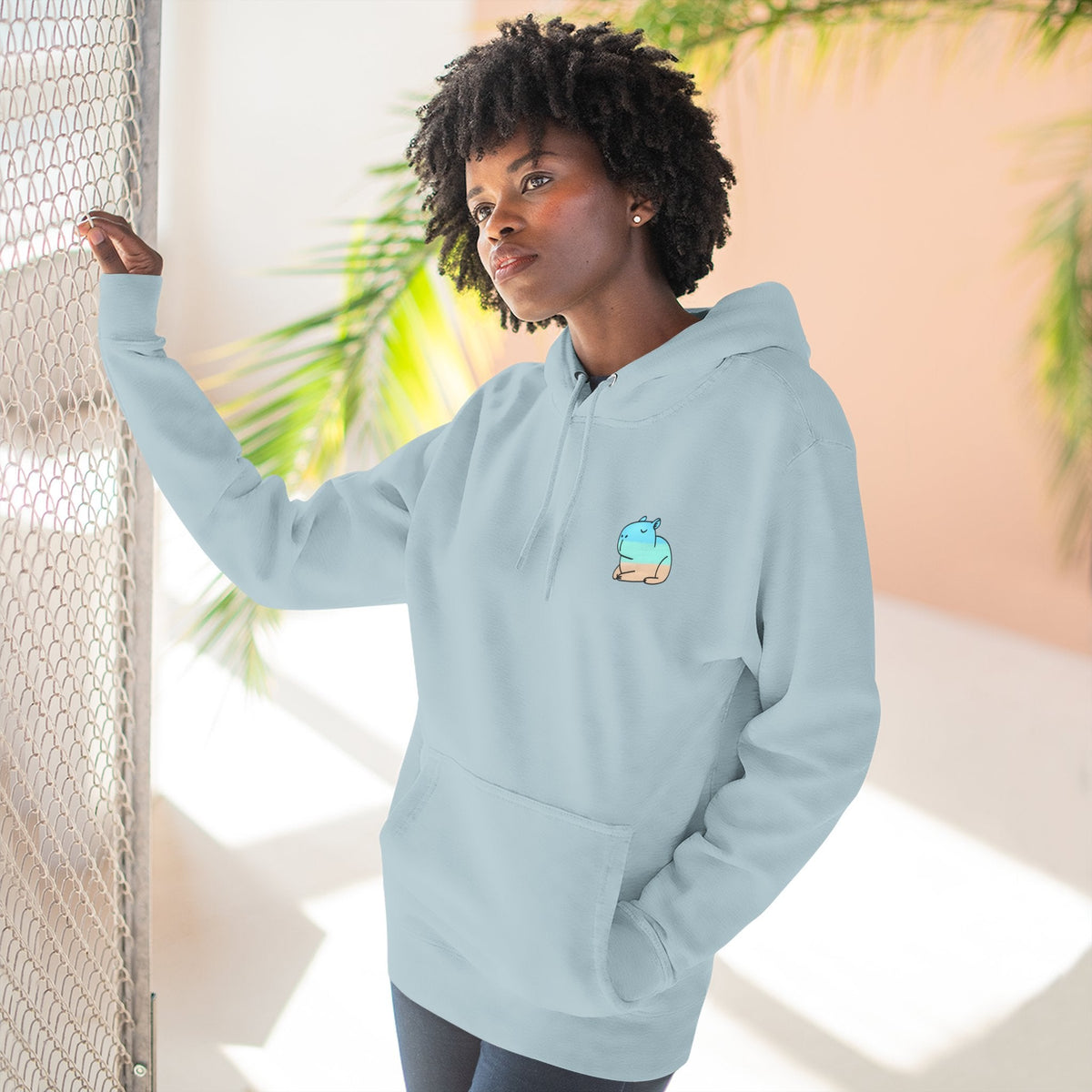 Capy Life coastal capybara hoodie in light blue with a small minimalist graphic, worn by a woman near a fence.