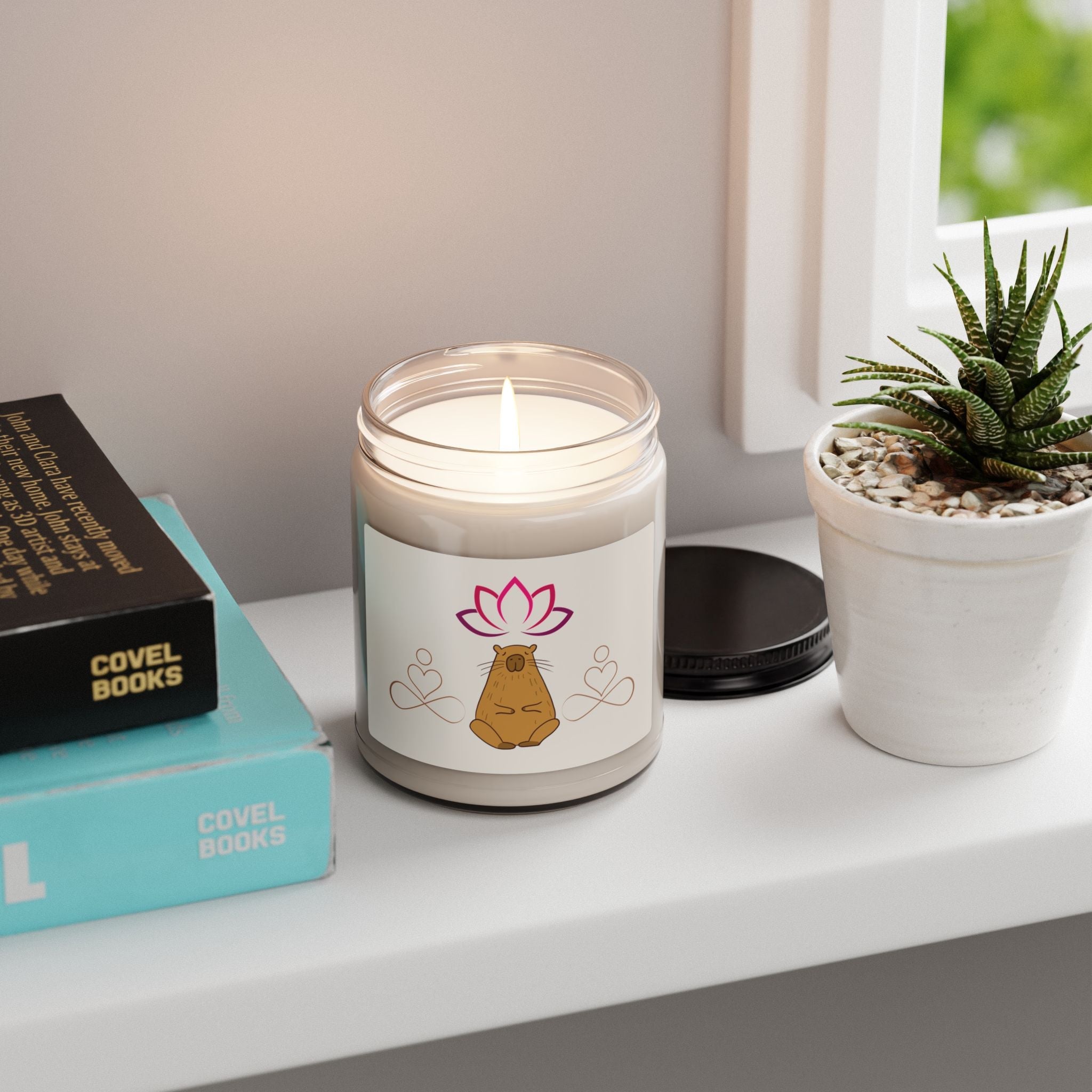 Capy Life scented candle in a glass jar with a meditating capybara and lotus label, sitting on a windowsill by books.