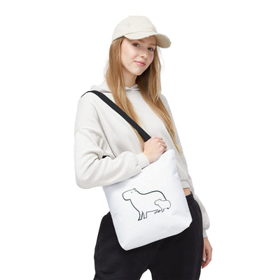 A woman wearing a beige hoodie and cap carries a white Capy Life minimalist tote bag with a capybara line art print.