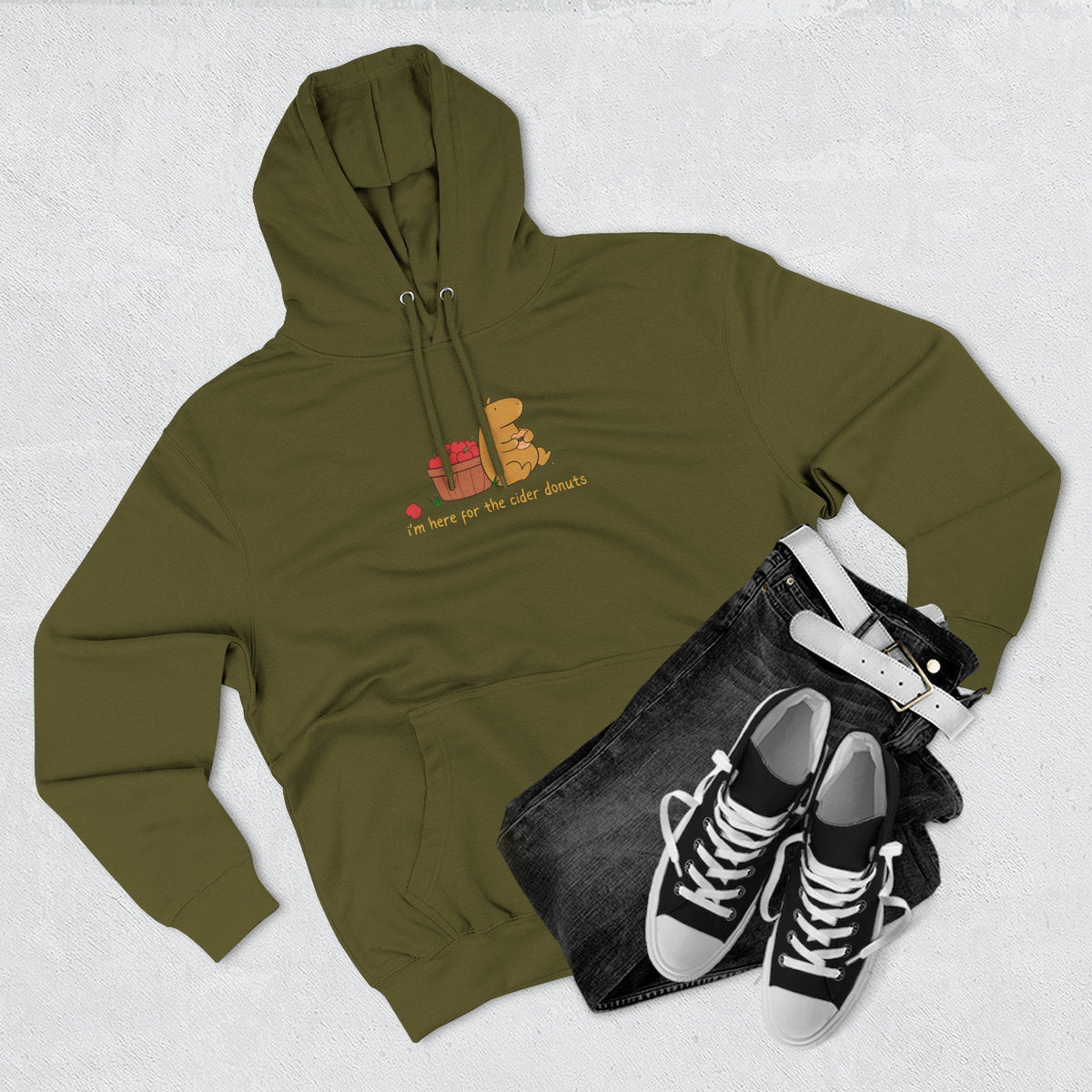 Capy Life olive green fleece hoodie featuring a capybara eating a donut, styled with black jeans and sneakers.