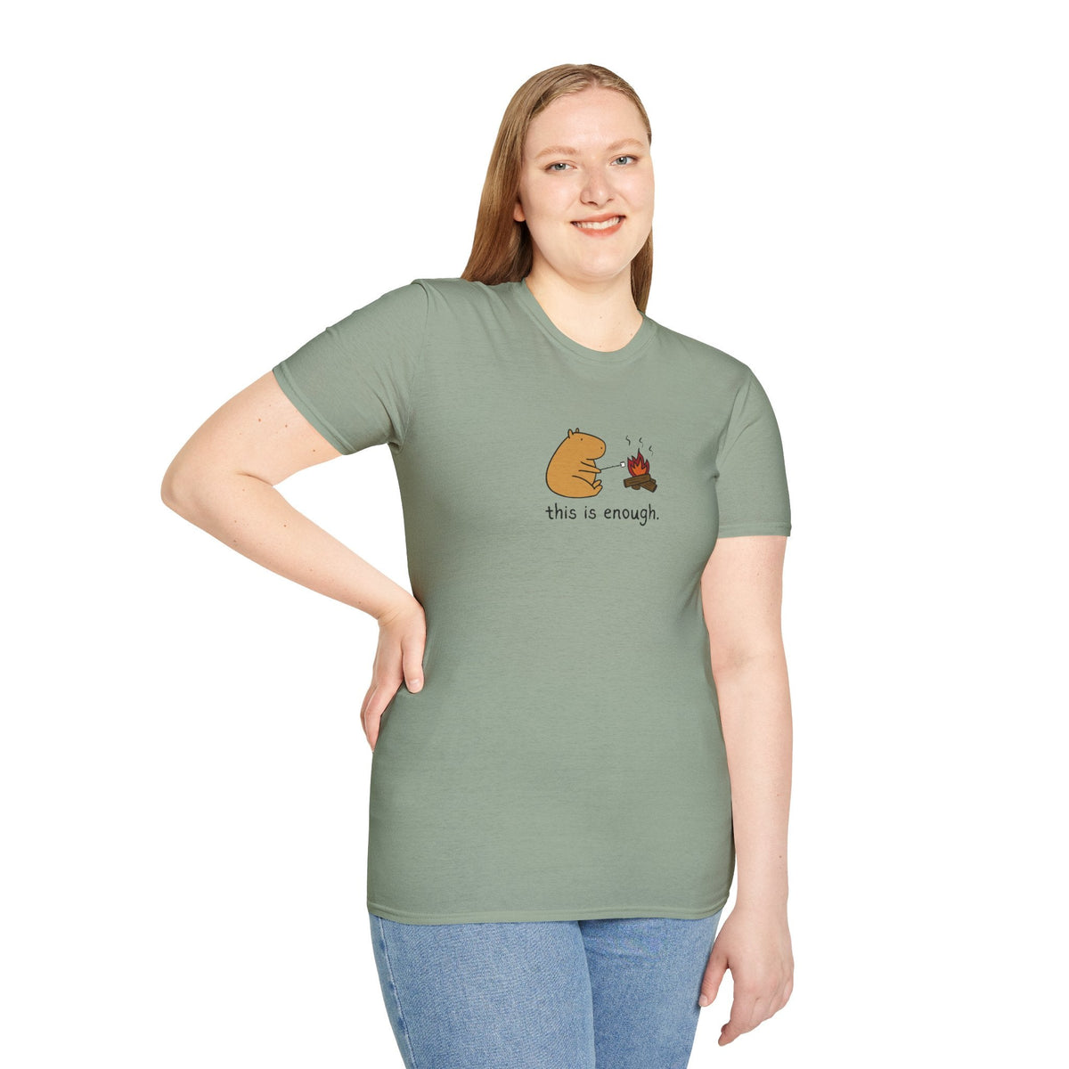 A woman wearing a sage green Capy Life softstyle t-shirt featuring a capybara roasting a marshmallow by a campfire.